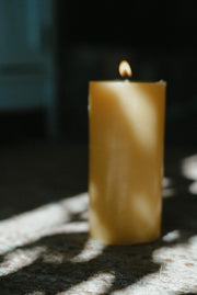 Lit Busy Bees 3 Inch Pillar Trio candle glowing beside a window with sunlight casting shadows across the surface.