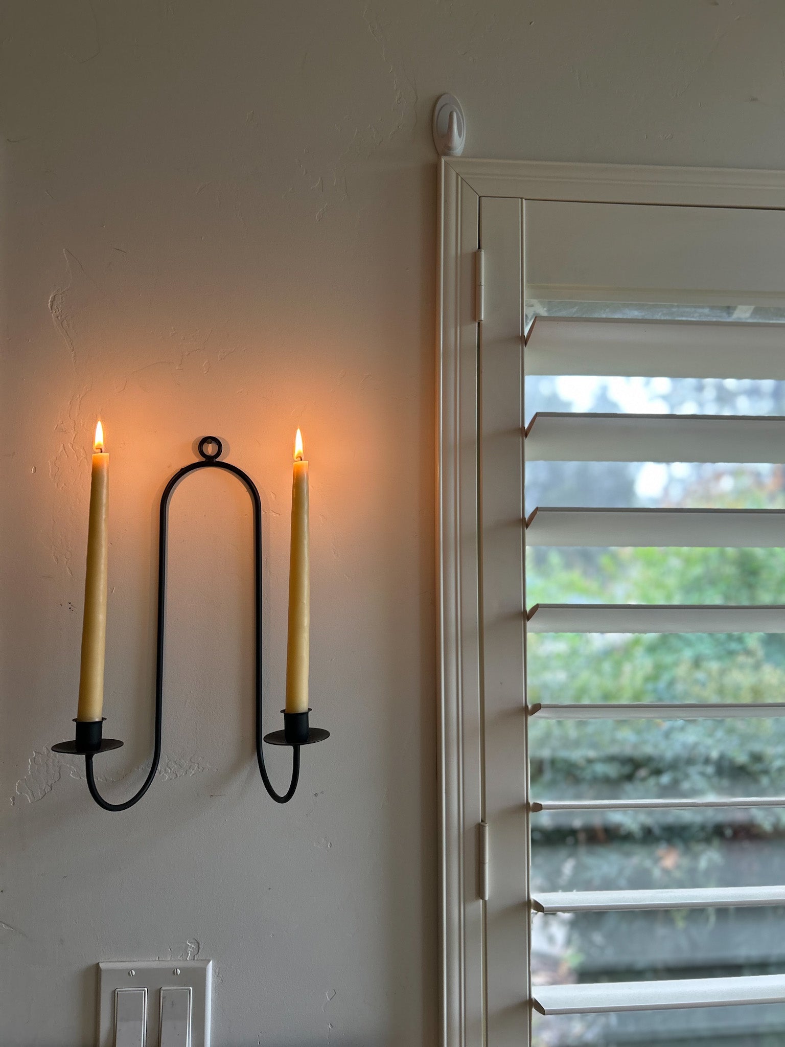 Lit Busy Bees Hand Dipped 12 Inch Taper Candles mounted on wall sconces beside a window in a cozy interior setting.