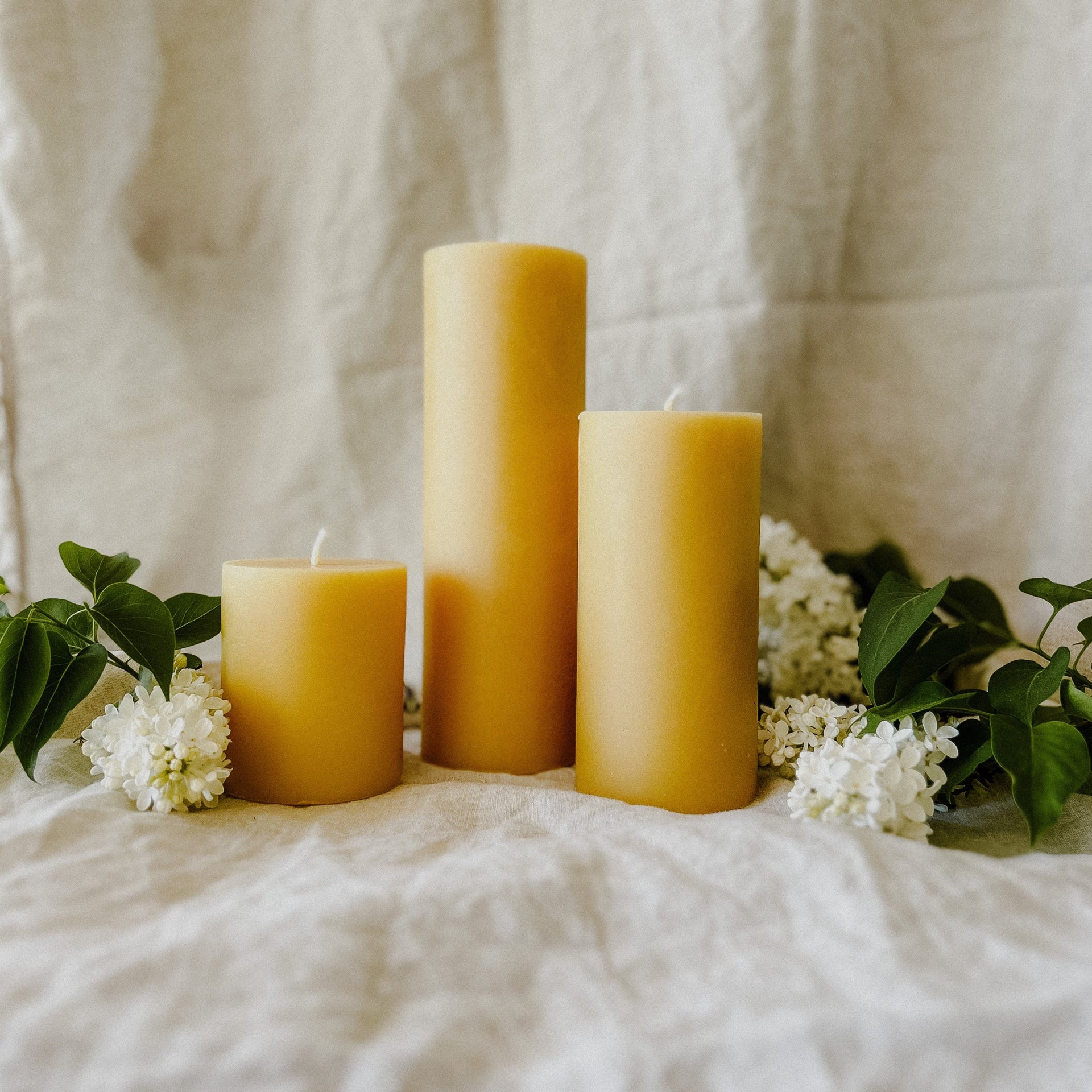 Busy Bees 3 Inch by 9 Inch pillar candles arranged on draped fabric with greenery accents in a warm minimal setting.