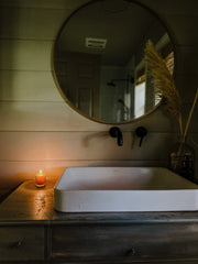 Lit Busy Bees Votive Candle from the 4 Pack glowing beside a bathtub and round mirror in a dimly lit bathroom.