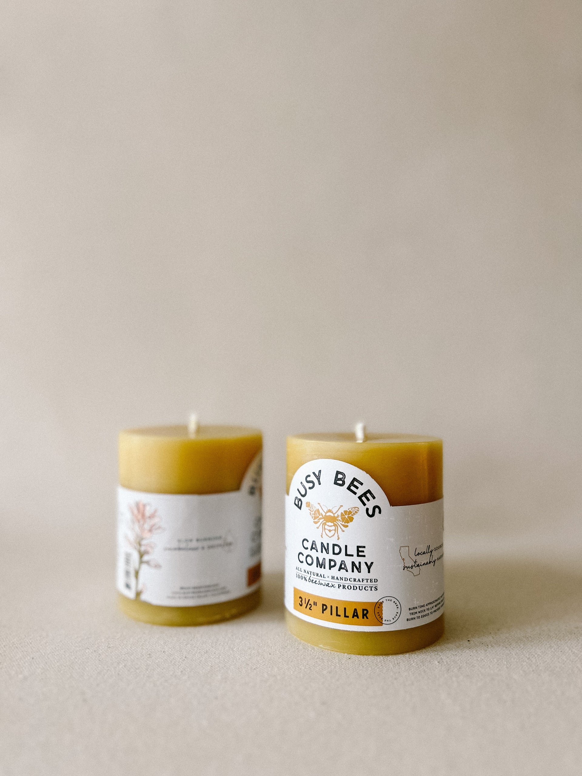 Two Busy Bees 3 Inch Pillar Trio beeswax candles with branded labels displayed on neutral fabric.