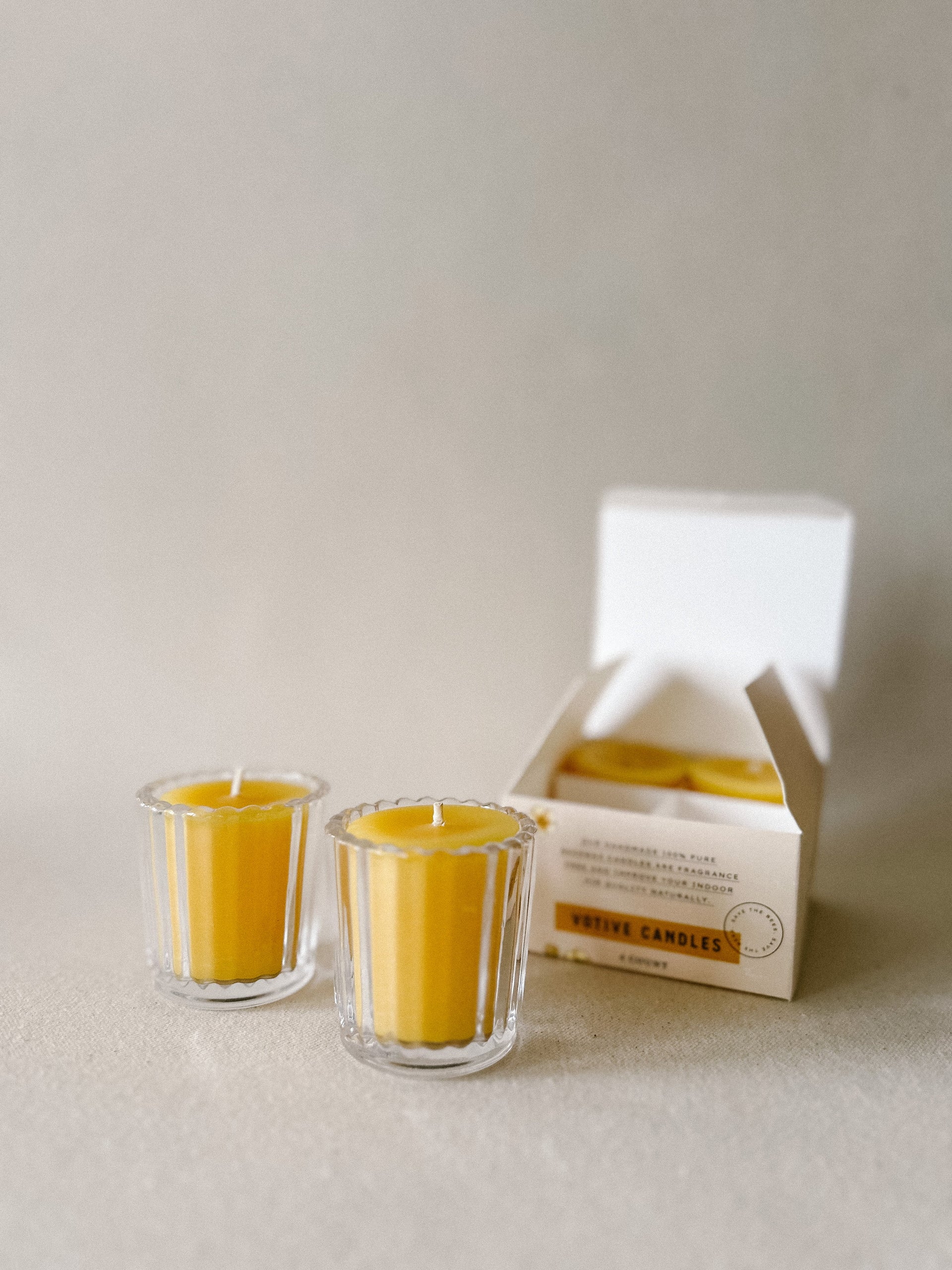 Two lit Busy Bees Votive Candles from the 4 Pack glowing against a soft neutral backdrop.