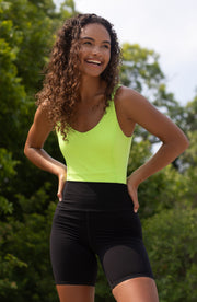 Woman smiling outdoors while wearing the Elastique Athletics Riviera neon green sleeveless top paired with black fitted shorts.
