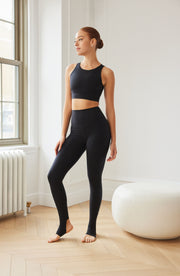 Woman standing indoors near a window while wearing the Elastique Athletics L'Original Lymphatic Stirrup Leggings in black with a matching sleeveless top.