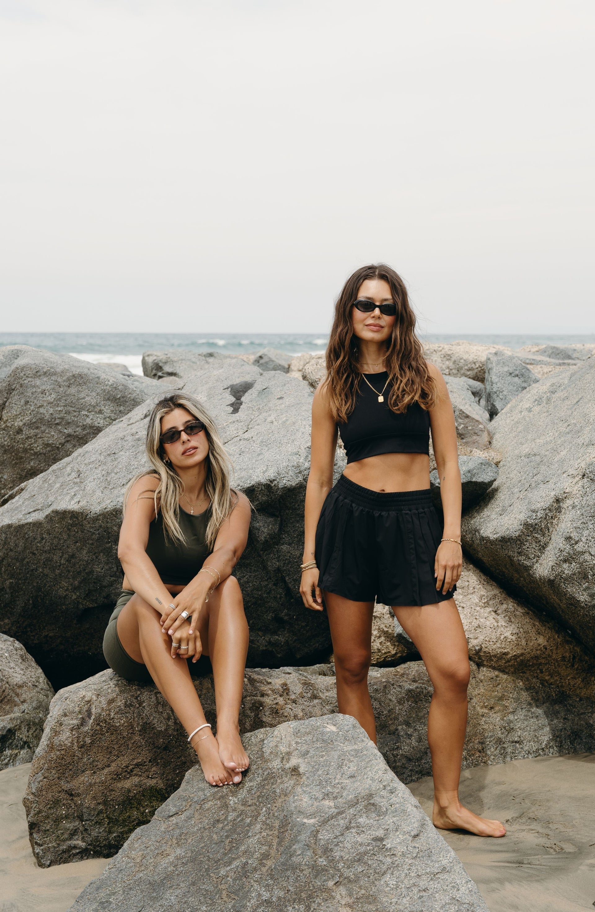 Two women sitting on coastal rocks while wearing the Elastique Athletics L'Original Flowy Short in black with matching athletic tops.