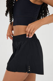 Close up side detail of the Elastique Athletics L'Original Flowy Short in black showing the lightweight layered fabric and curved hem.