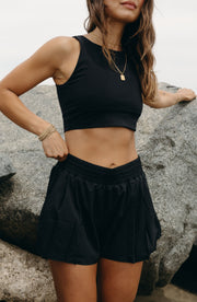 Woman posing outdoors on rocks while wearing the Elastique Athletics L'Original Flowy Short in black with a matching sports bra.