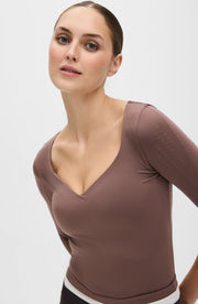 Close up portrait of a woman wearing the Elastique Athletics Iconic Sleeve MicroPerle Top in taupe with a deep scoop neckline and fitted long sleeves.