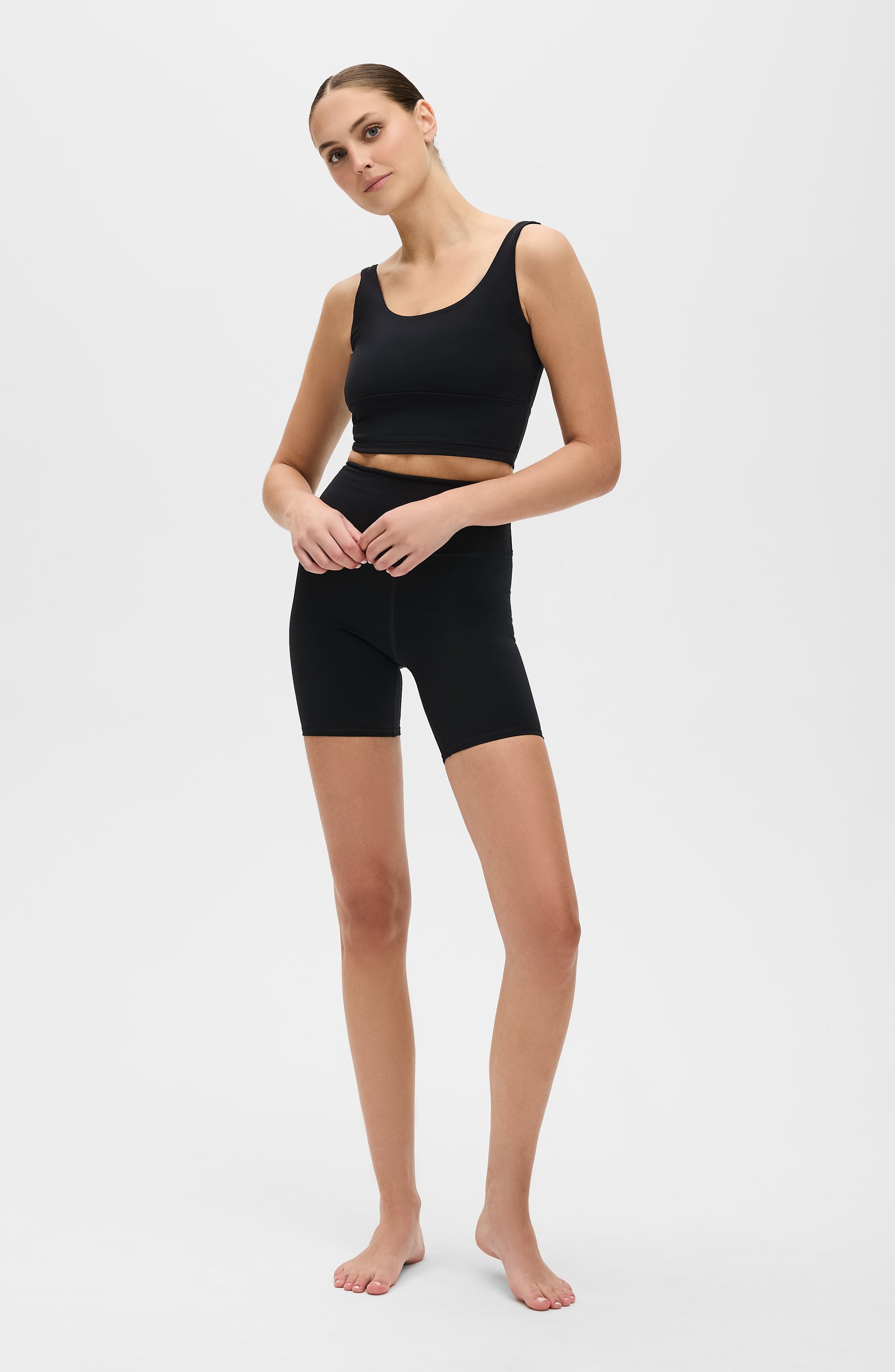 Woman standing barefoot in a studio while wearing the Elastique Athletics Lisse black sleeveless crop top and matching fitted biker shorts.