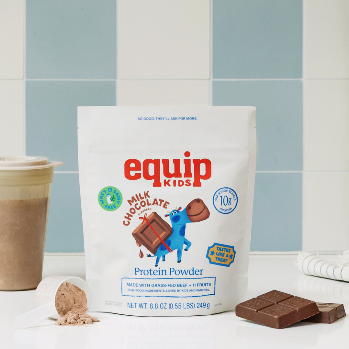 Equip Kids Milk Chocolate Protein Powder pouch with a chocolate shake, chocolate pieces, and a scoop of powder on a blue tiled backdrop.