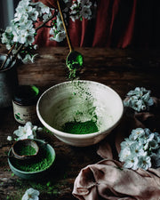 Ceremonial grade matcha powder cascading from a spoon into a handcrafted bowl surrounded by white blossoms and rustic wood accents.