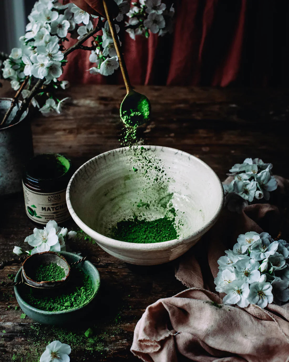 Ceremonial grade matcha powder cascading from a spoon into a handcrafted bowl surrounded by white blossoms and rustic wood accents.
