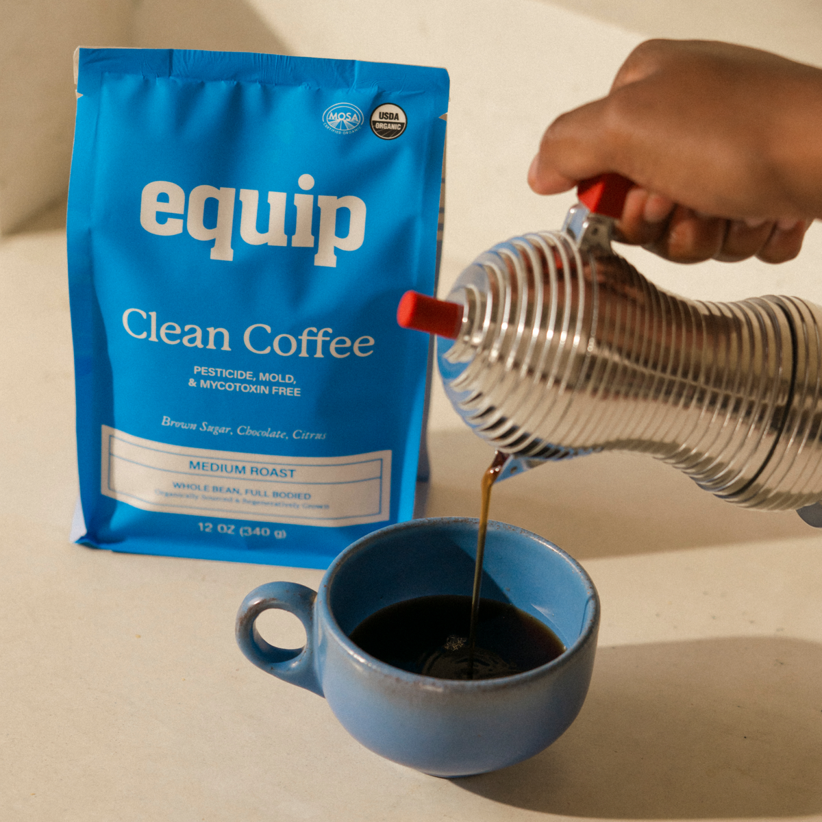 Equip Clean Coffee medium-roast caffeinated coffee bag beside a blue cup as coffee is poured from a metal carafe.