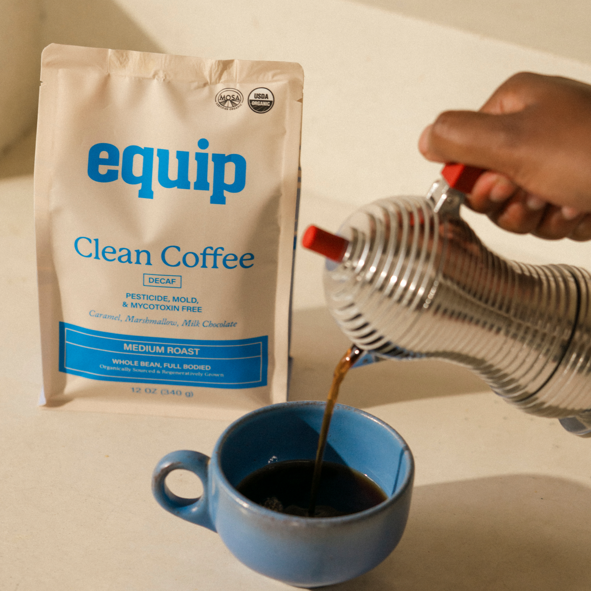 Equip Clean Coffee medium-roast decaf bag beside a blue cup as coffee is poured from a metal carafe.