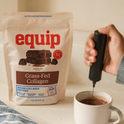 Equip Grass-Fed Collagen chocolate pouch beside a white mug as a handheld frother mixes a chocolate drink.