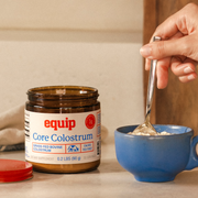 Open Equip Core Colostrum jar beside a blue cup while colostrum powder is stirred.