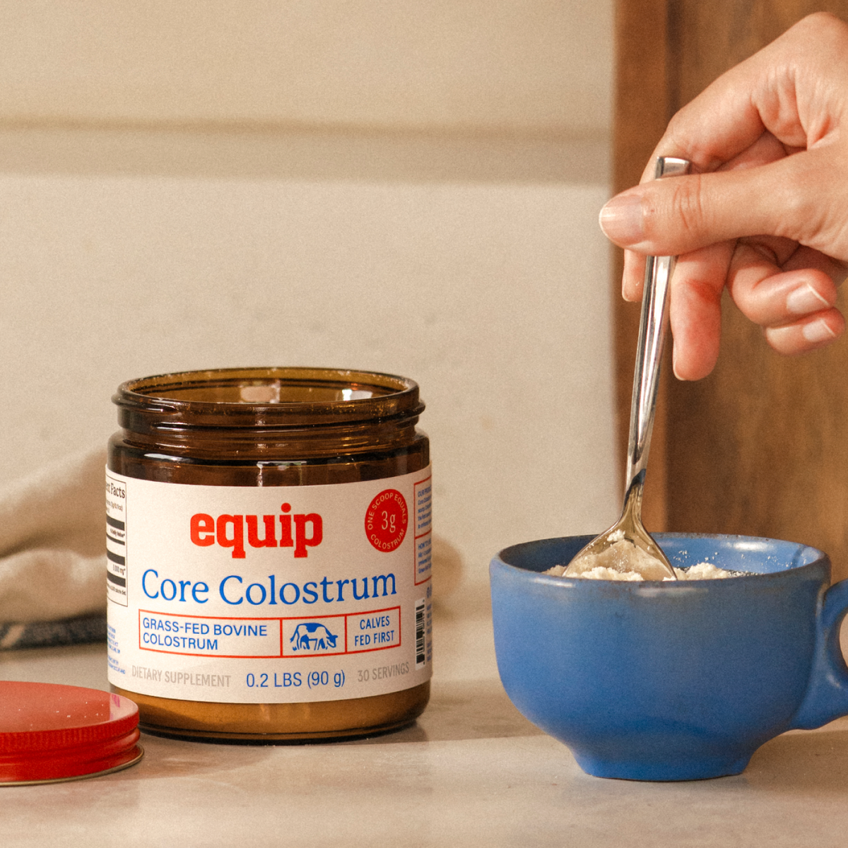 Open Equip Core Colostrum jar beside a blue cup while colostrum powder is stirred.