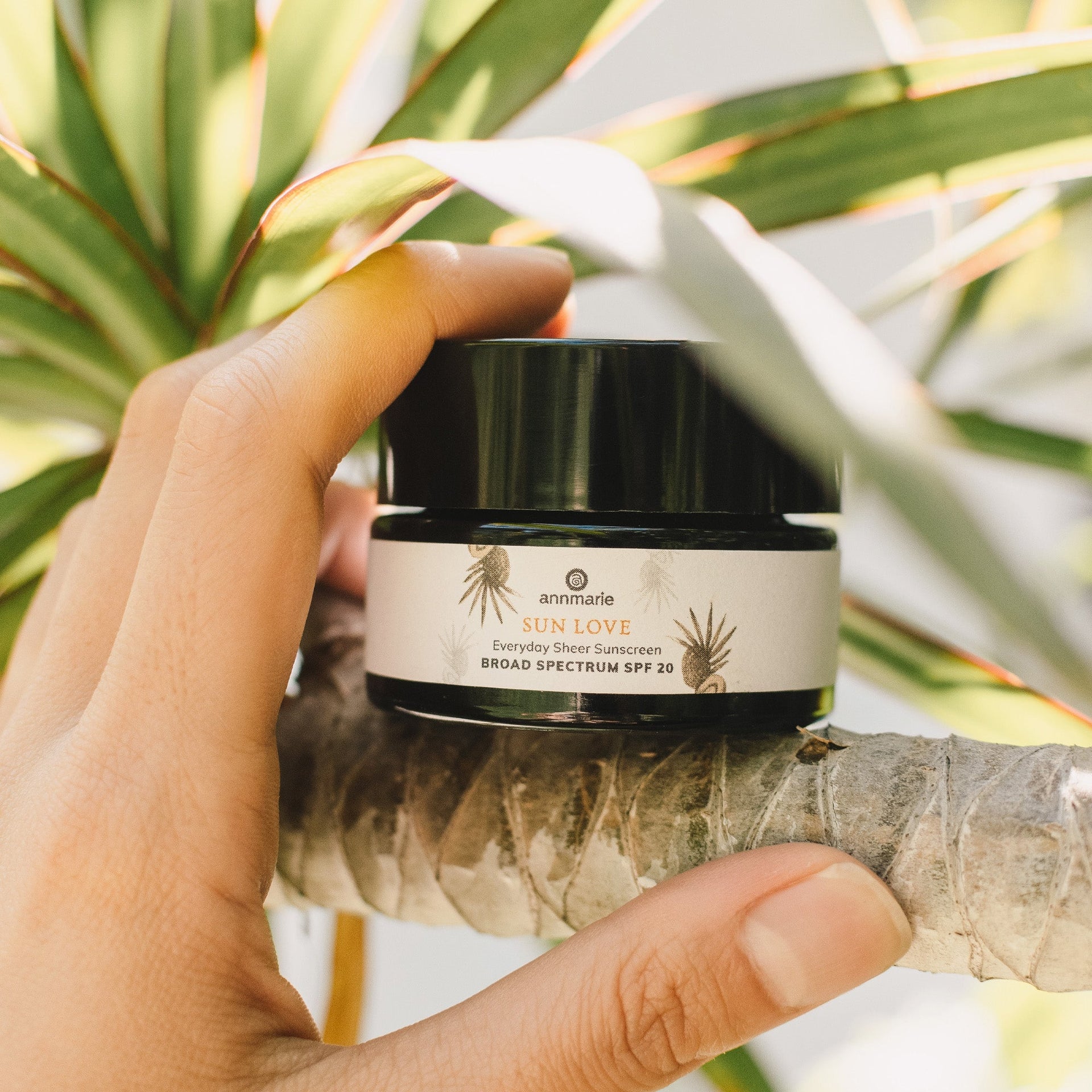 Annmarie Deluxe Mini Sun Love travel-size mineral sunscreen held outdoors among tropical foliage