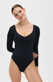 Woman wearing the Elastique Athletics Iconic long sleeve black bodysuit poses with her head tilted and hands behind her back against a light neutral background.