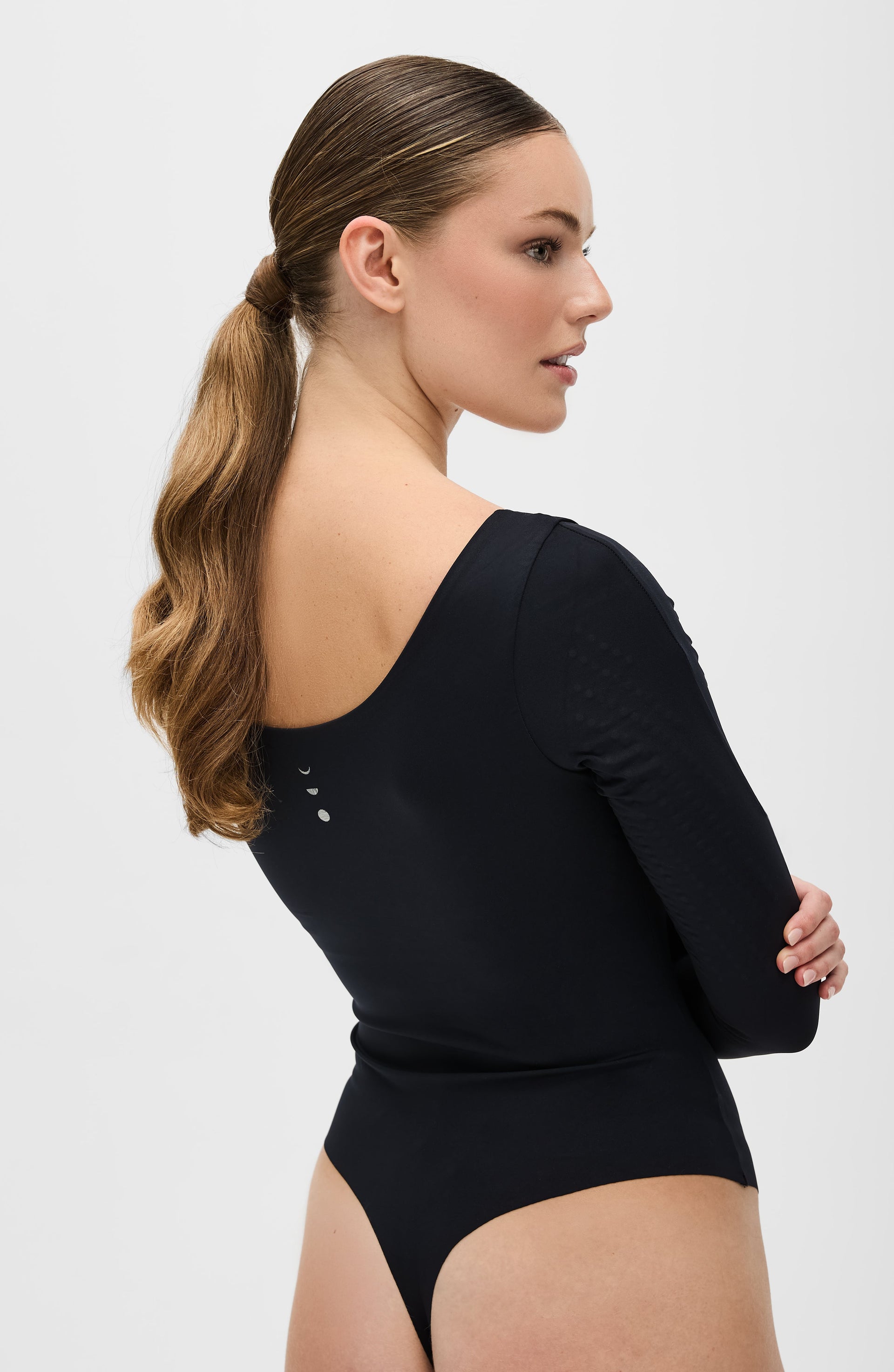 Rear three quarter view of a woman wearing the Elastique Athletics Iconic long sleeve black bodysuit with her hair tied back in a ponytail.
