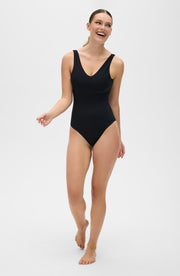 Woman walking barefoot in a studio while wearing the Elastique Athletics Adorn black sleeveless bodysuit with a deep scoop neckline.