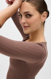 Close up side portrait of a woman wearing the Elastique Athletics Iconic Sleeve MicroPerle Top in taupe with one arm raised beside her face.