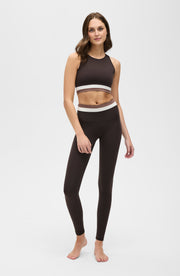 Woman standing barefoot in a studio while wearing the Elastique Athletics Le Monde black sleeveless crop top with white trim and matching high waisted leggings.