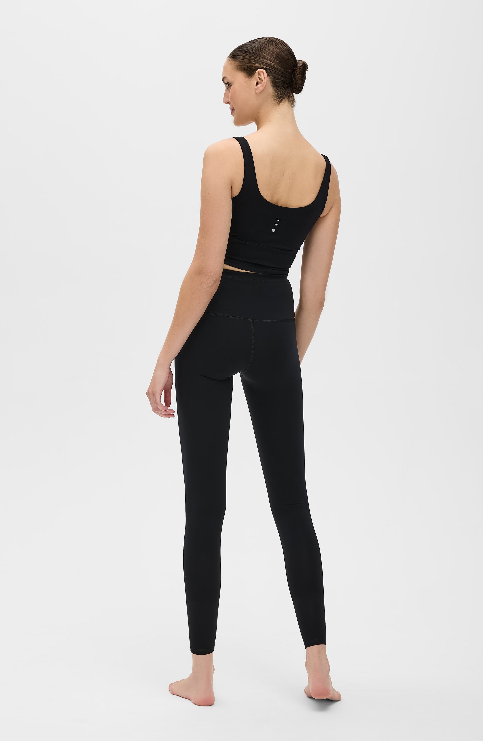 Back view of a woman standing barefoot while wearing the Elastique Athletics Lisse Compression Bra in black with matching leggings.