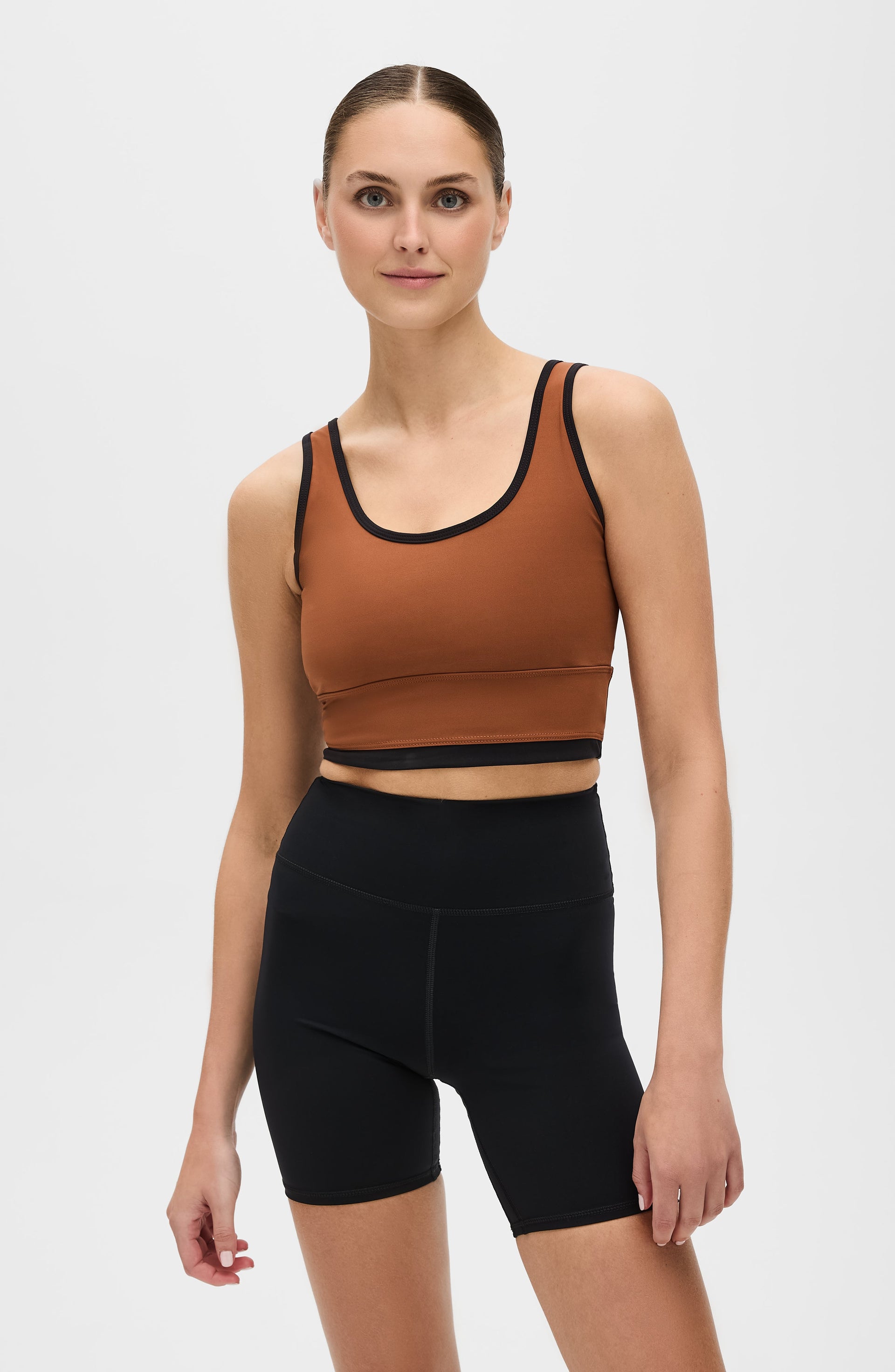 Woman standing barefoot while wearing the Elastique Athletics Lisse Bra in chestnut brown with black trim and matching black biker shorts.