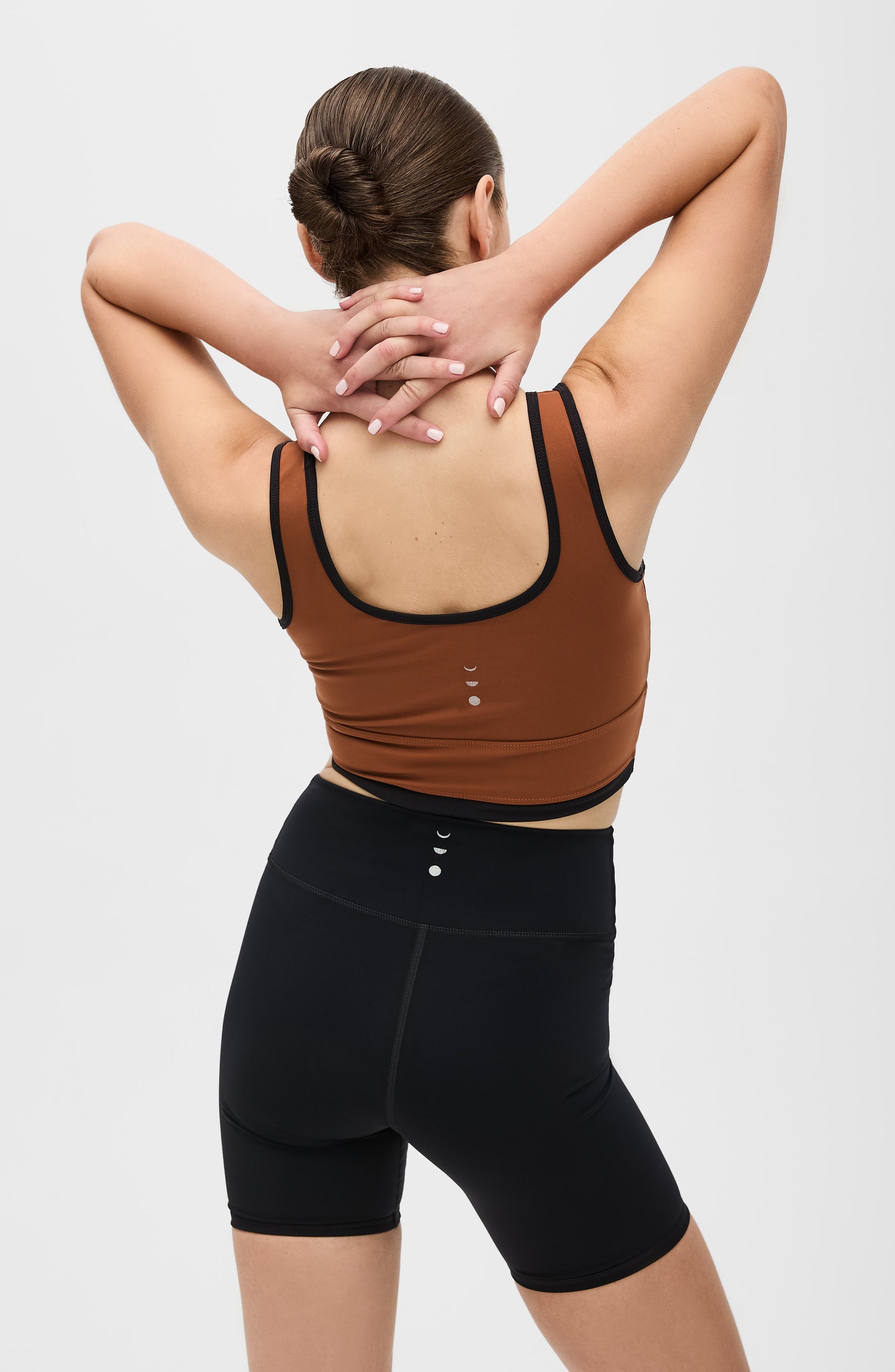 Back view of a woman with both hands behind her neck while wearing the Elastique Athletics Lisse Bra in chestnut brown with black biker shorts.