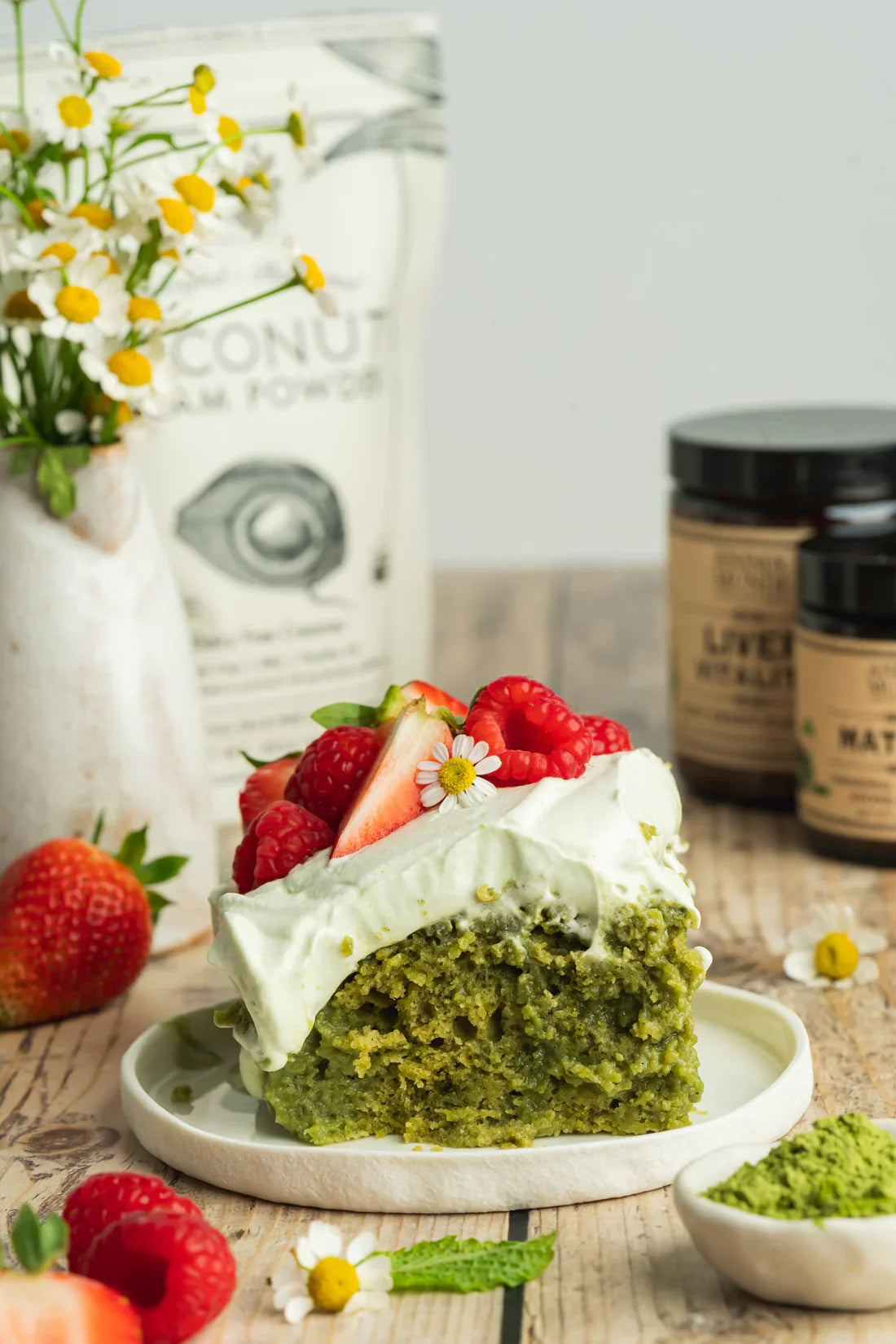 Slice of matcha cake topped with whipped cream, berries, and fresh flowers beside ceremonial matcha powder.