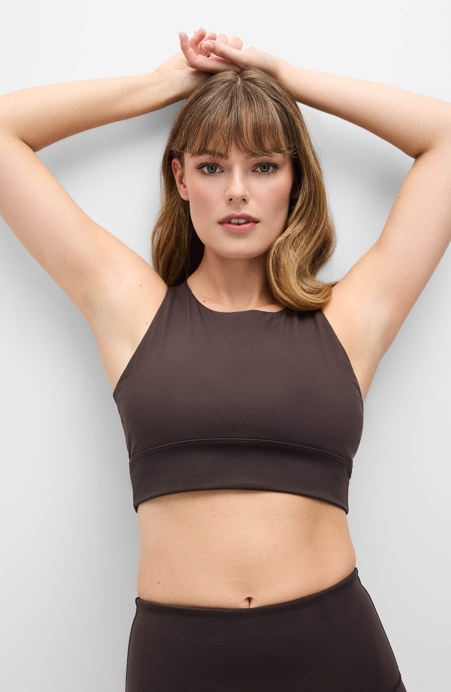 Woman posing with both hands above her head while wearing the Elastique Athletics L'Original dark brown sports bra and matching leggings.