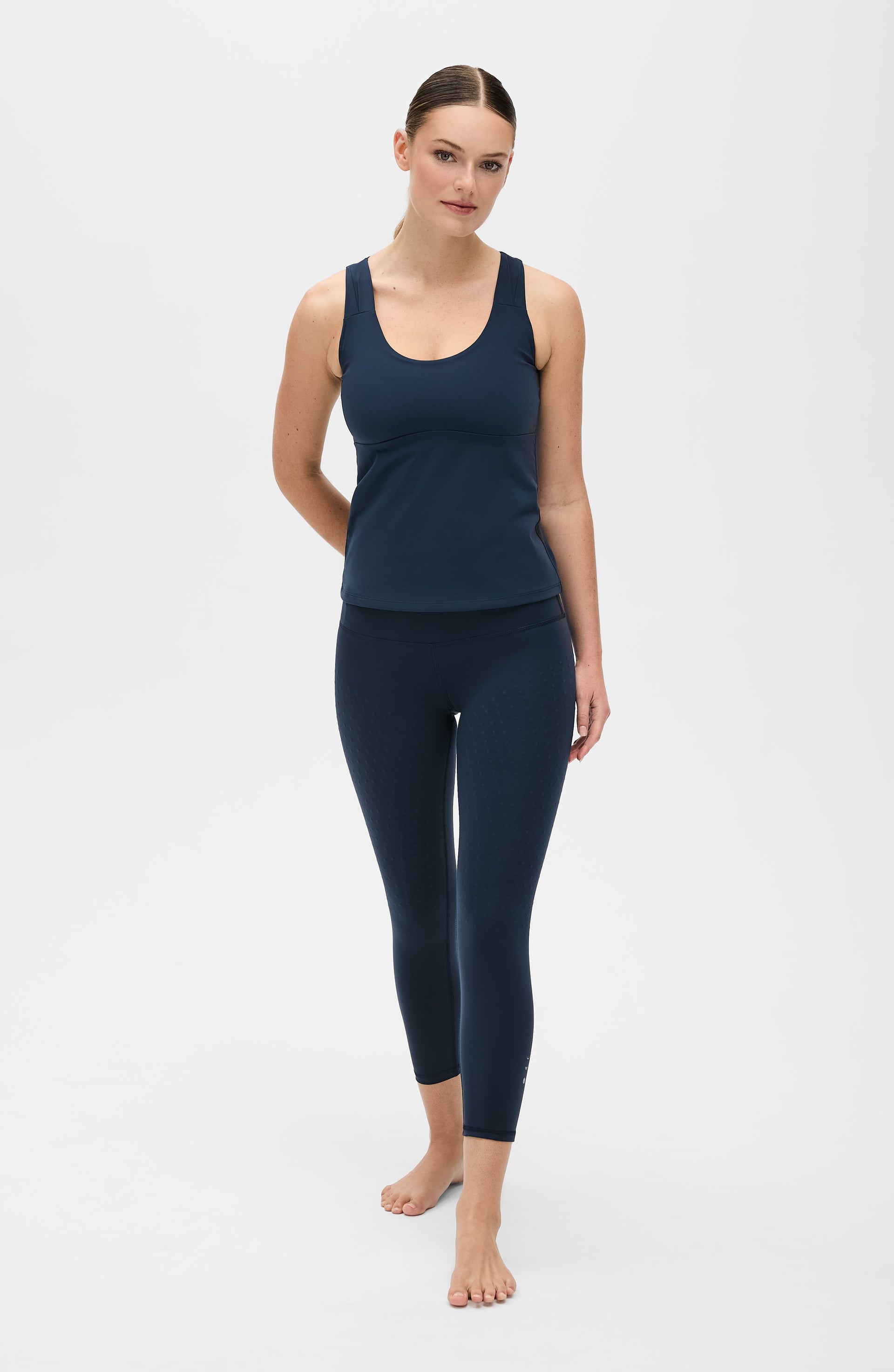 Full body studio portrait of a woman walking barefoot while wearing the Elastique Athletics L'Original Lymphatic Tank in navy blue with matching leggings.