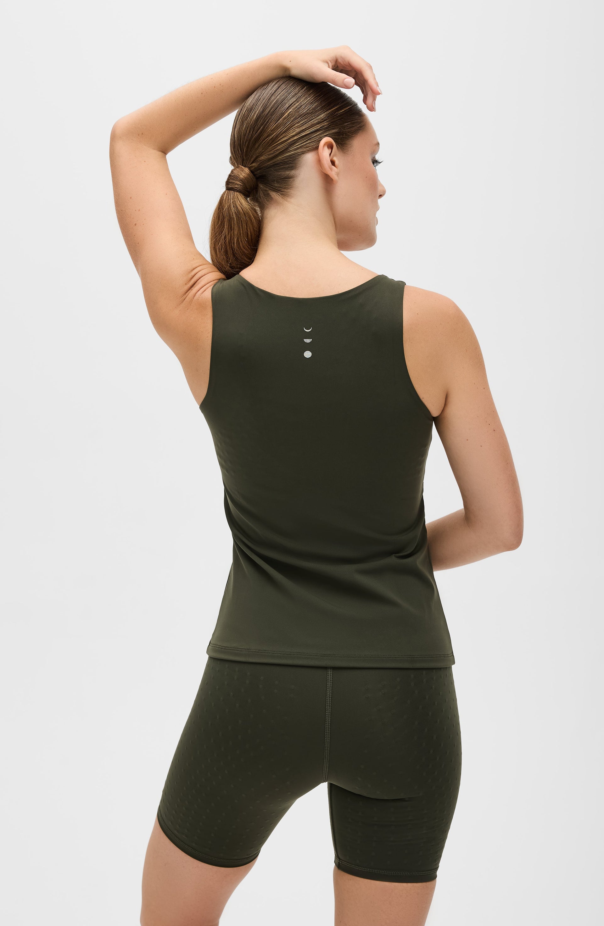 Woman posing with one arm raised while wearing the Elastique Athletics L'Original Lymphatic Tank in olive green with matching bike shorts.