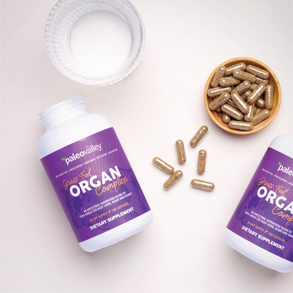 Paleovalley Grass Fed Organ Complex bottles surrounded by capsules and a glass of water, showcasing a nutrient-dense organ supplement formula.