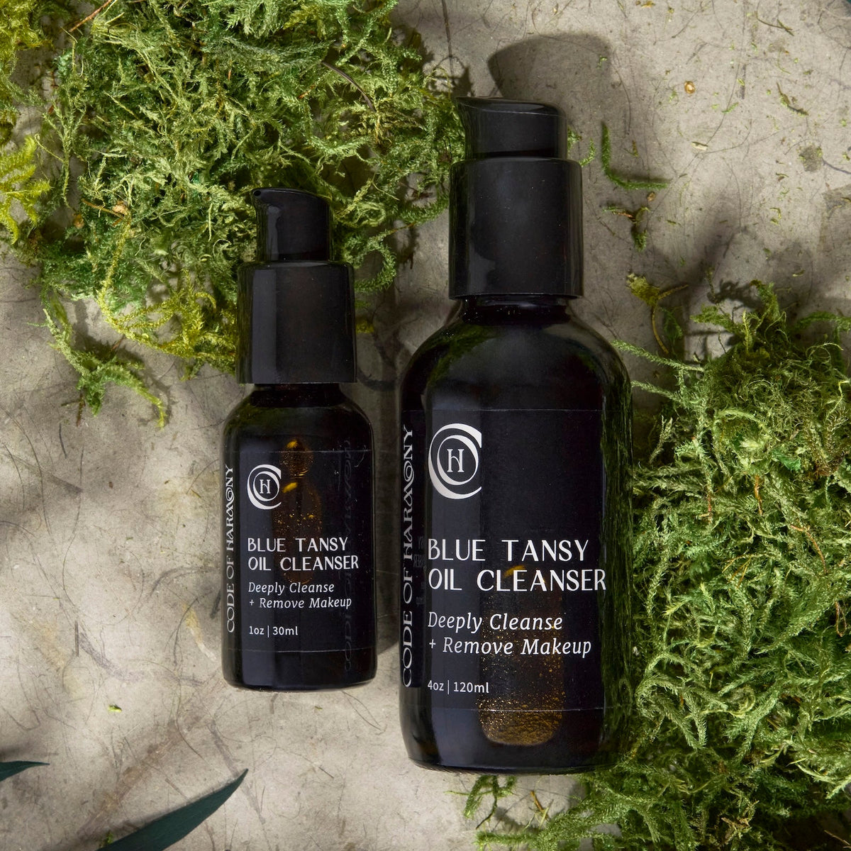 Code of Harmony Blue Tansy Oil Cleanser 1 oz and 4 oz bottles styled with green moss