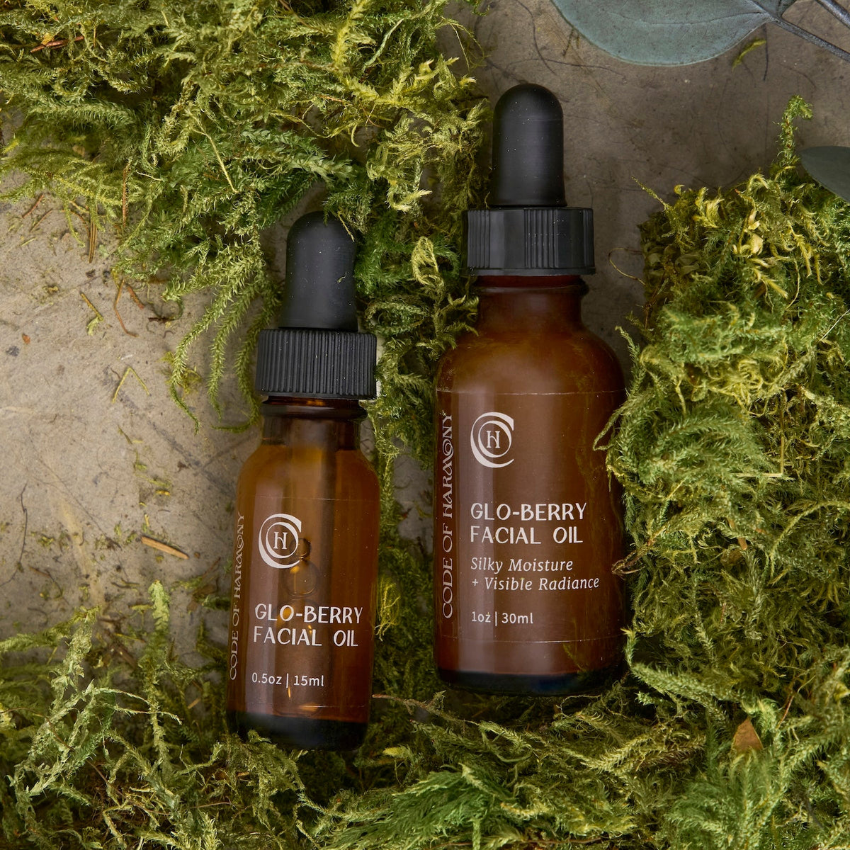 Code of Harmony Glo-Berry Facial Oil 0.5 oz and 1 oz bottles styled with green moss