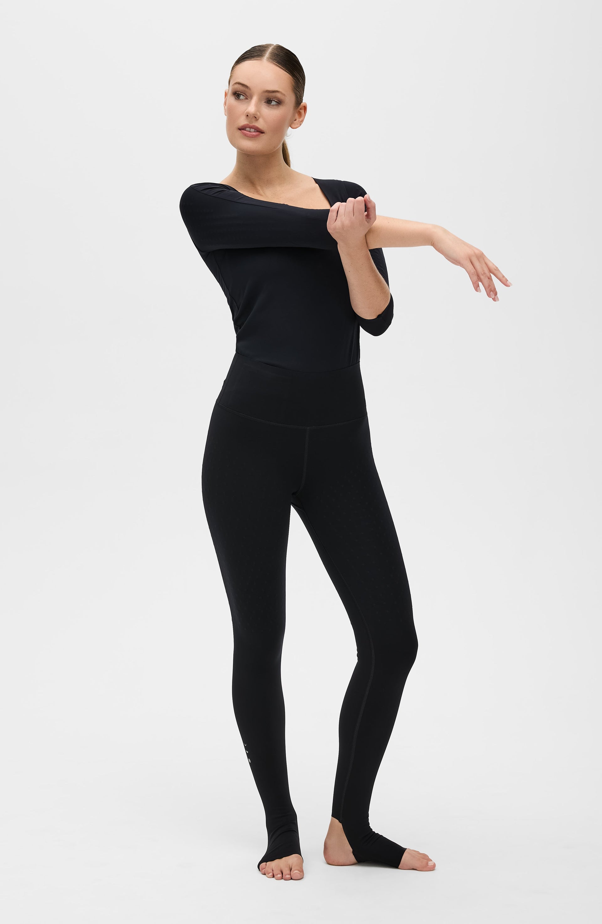 Woman posing in a studio while wearing the Elastique Athletics L'Original Lymphatic Stirrup Leggings in black with a fitted tank top.
