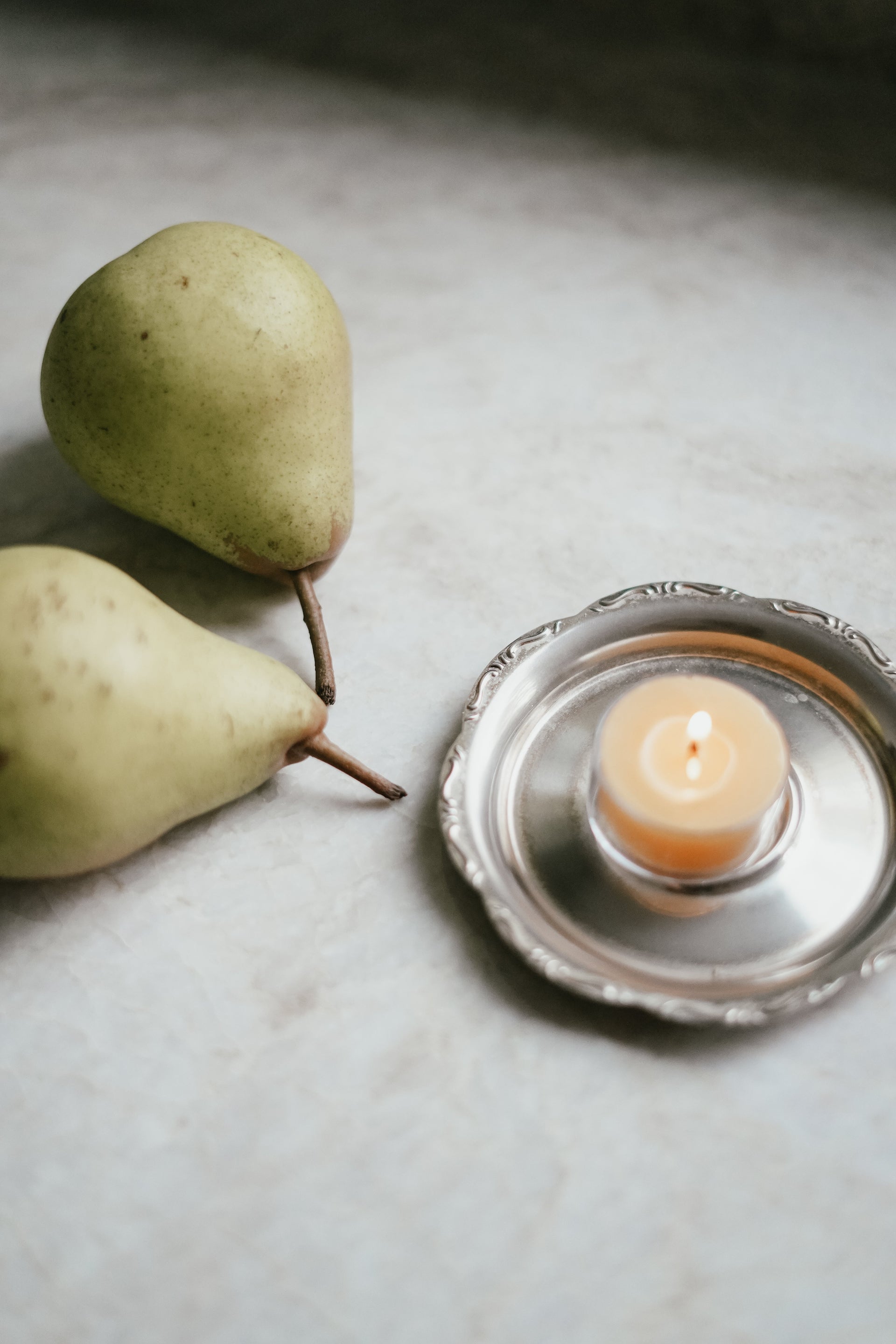Busy Bees Bulk Loose Tea Light Candle burning in a ribbed glass holder beside green pears on a neutral surface.