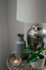 Busy Bees tea light candle from The Queen Bee Bundle glowing in a ribbed glass holder beside green glass bottles and flowers.